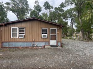 Small brown motel building with three windows and a door, surrounded by trees and gravel.